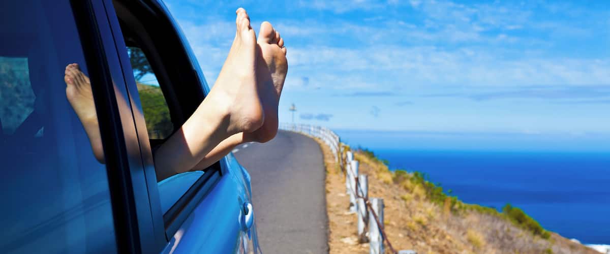 A car's foot resting on the window sill with a coastal road and ocean in the background.