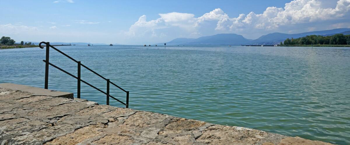 A stone pier with metal railings leading into a calm lake, surrounded by mountains and a clear sky.
