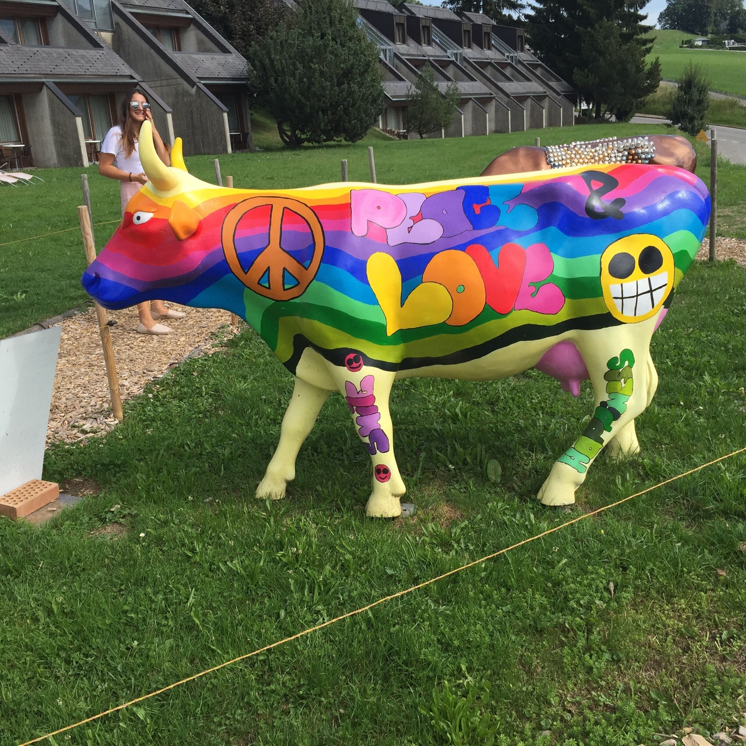 A colorful cow sculpture with peace and love symbols, set in a grassy area with houses in the background.