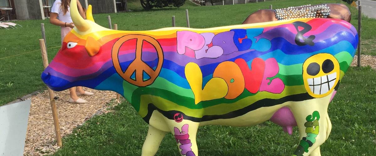 A colorful cow sculpture with peace and love symbols, set in a grassy area with houses in the background.