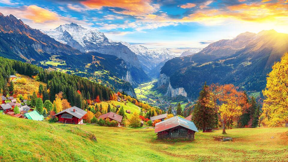 A mountainous landscape with a valley, houses, and autumn-colored trees.