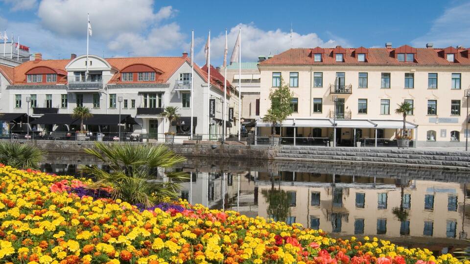A waterfront promenade with colorful flowers, a canal, and buildings with red-tiled roofs.
