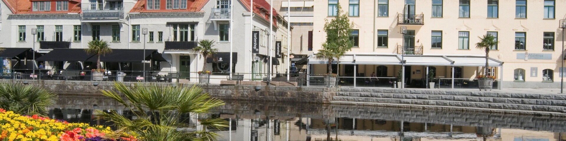 A waterfront promenade with colorful flowers, a canal, and buildings with red-tiled roofs.