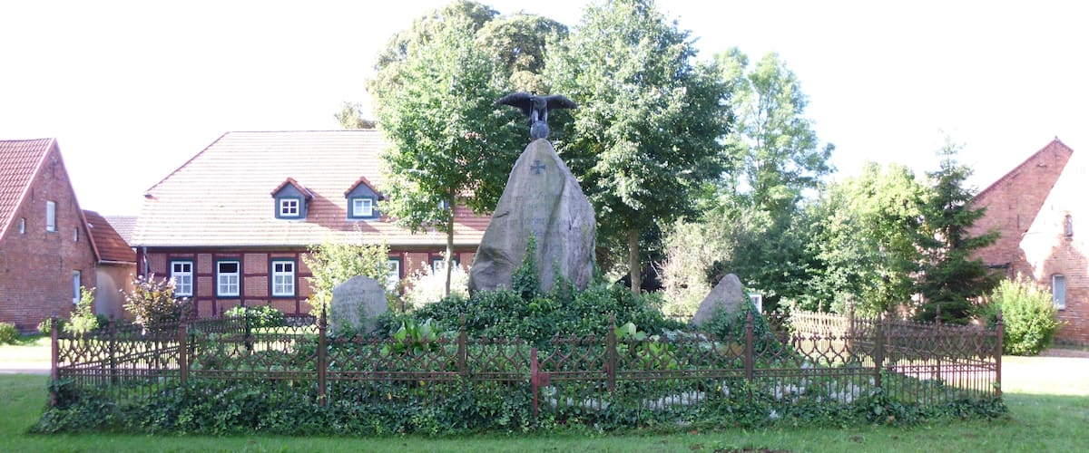 A garden with a large rock formation, a wooden fence, and a building with a red roof in the background.