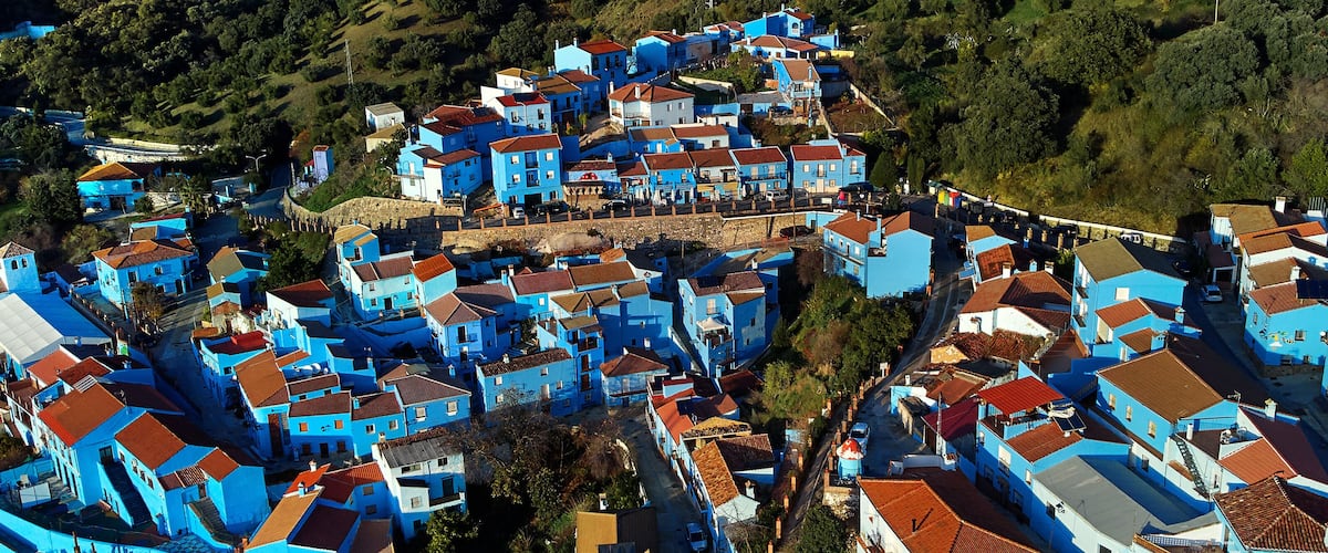 A densely packed village with blue buildings and red-tiled roofs, surrounded by greenery and hills.