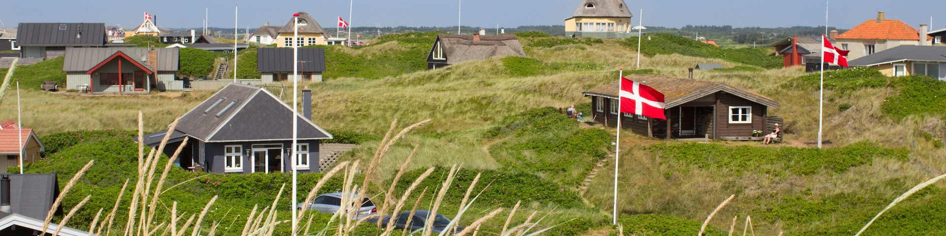 Un pueblo costero con casas sobre dunas cubiertas de hierba, un molino de viento y una bandera.