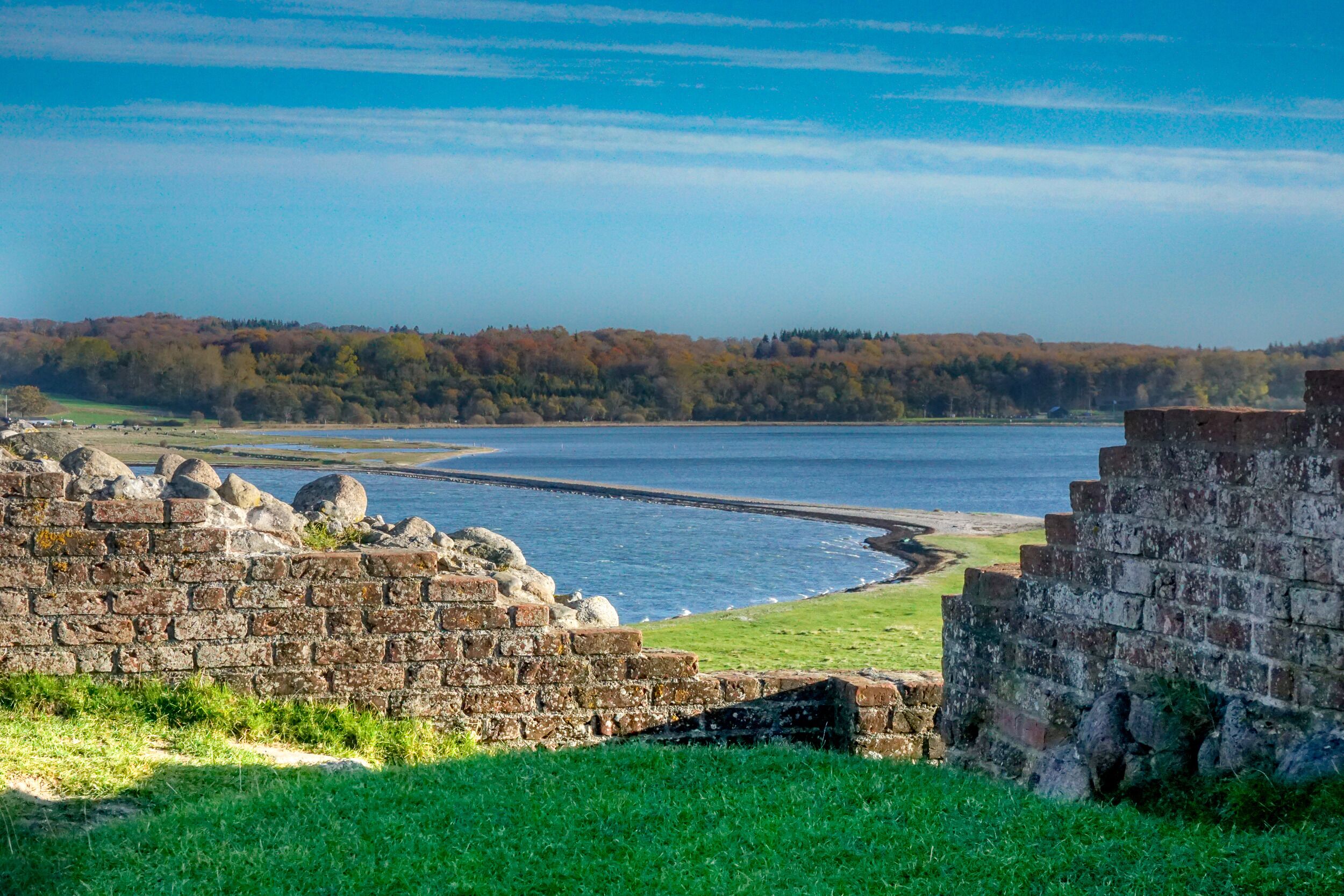 A historic stone wall by a lake with a forest in the background.