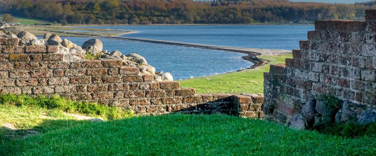 A historic stone wall by a lake with a forest in the background.
