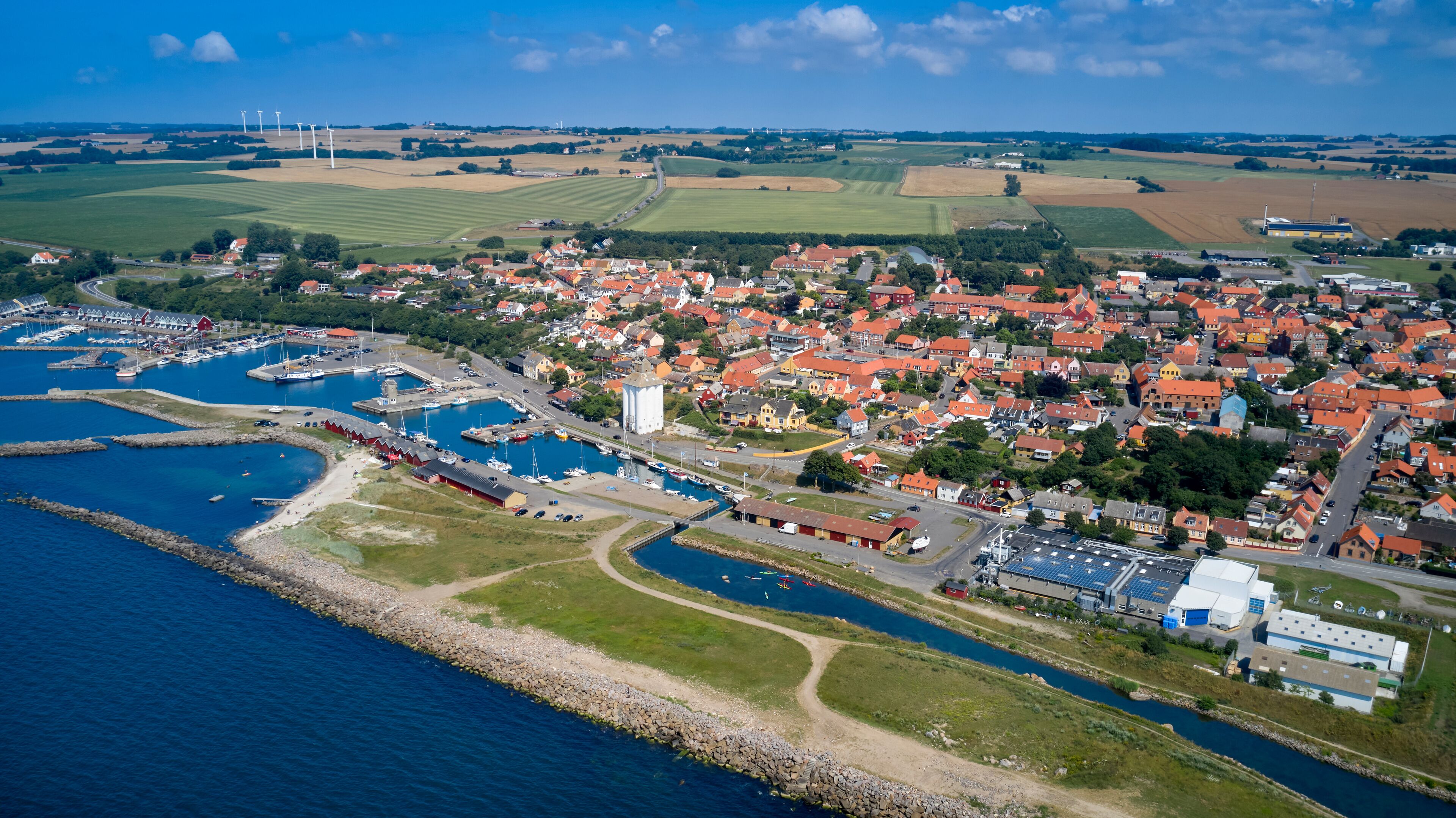 An aerial view of a coastal town with a marina, wind turbines, and agricultural fields.