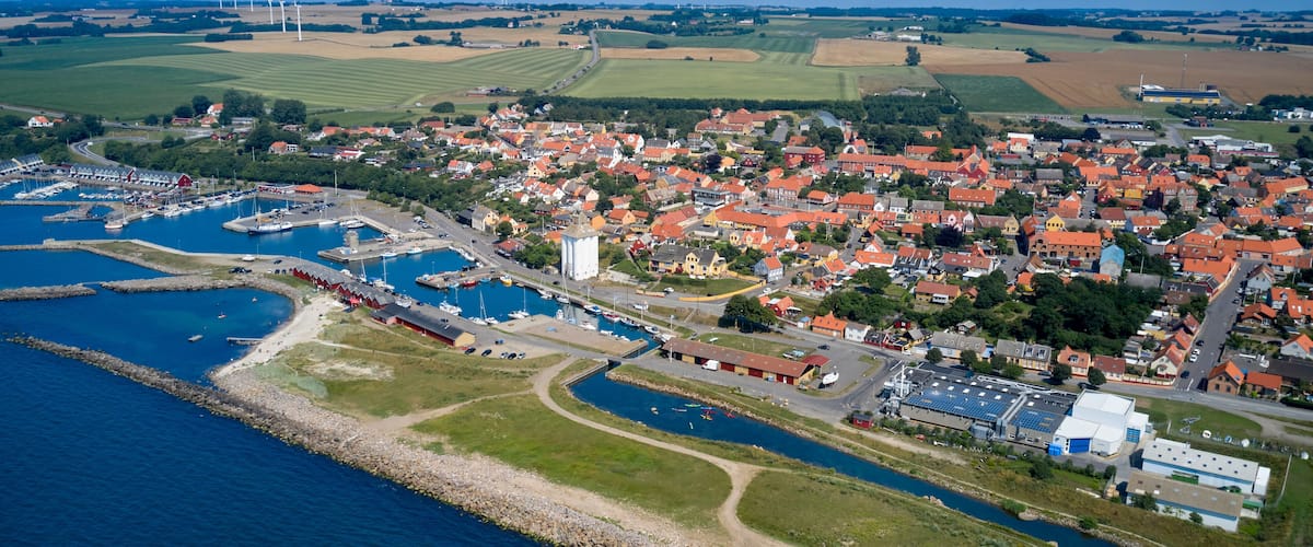 An aerial view of a coastal town with a marina, wind turbines, and agricultural fields.