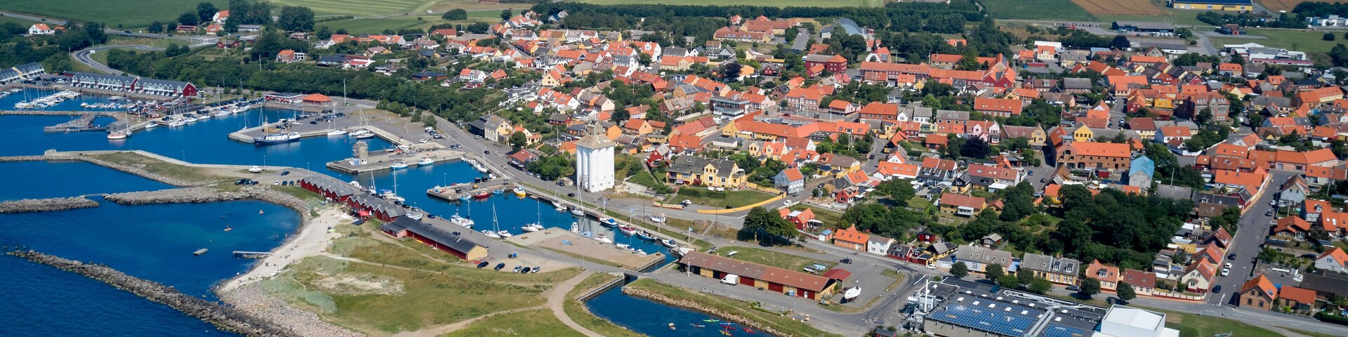An aerial view of a coastal town with a marina, wind turbines, and agricultural fields.