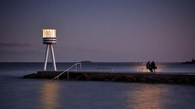 A calm sea at dusk with a lighthouse and a bench overlooking the water.