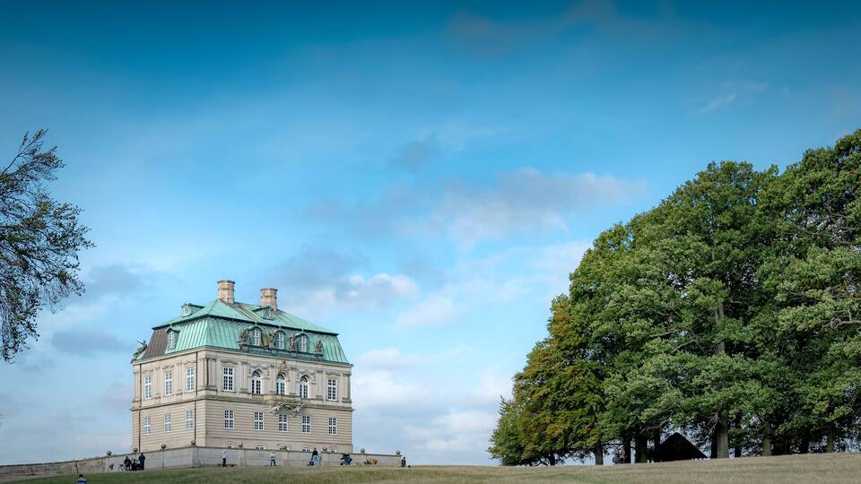 A historic building with a green roof and classical architecture, surrounded by trees and open grassland.