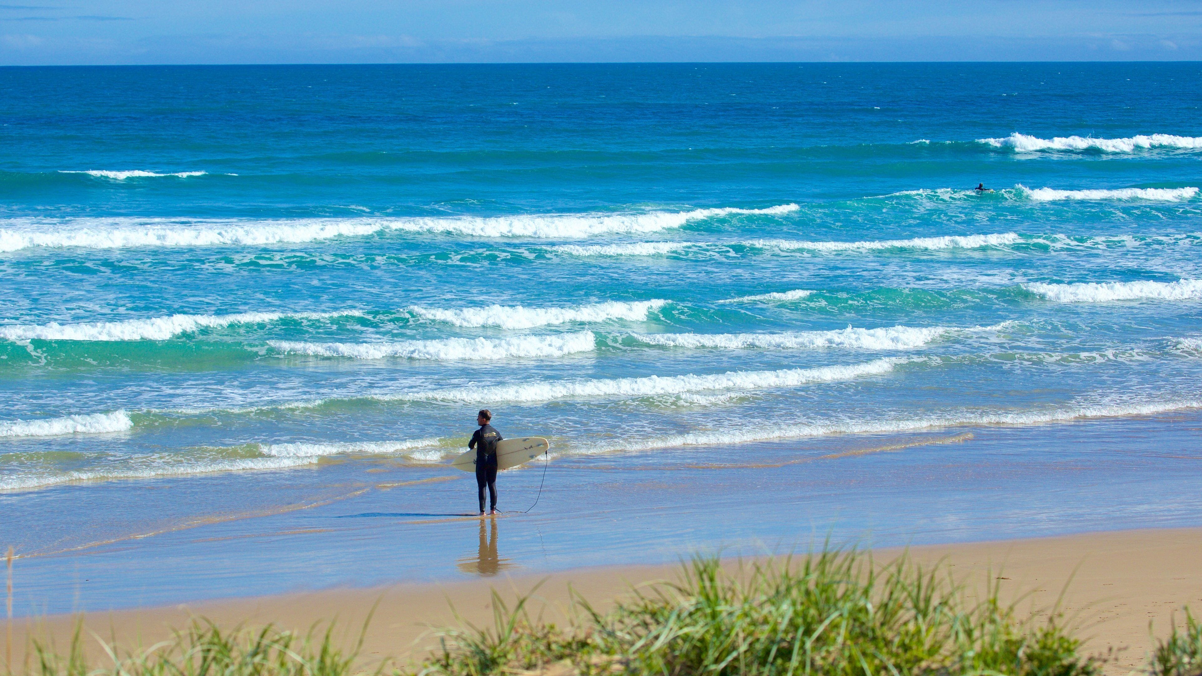 A surfer standing on a beach with waves approaching.