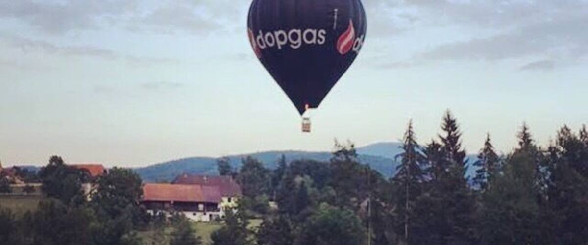 A hot air balloon with the word "dopgas" on it flying over a body of water with trees and buildings in the background.