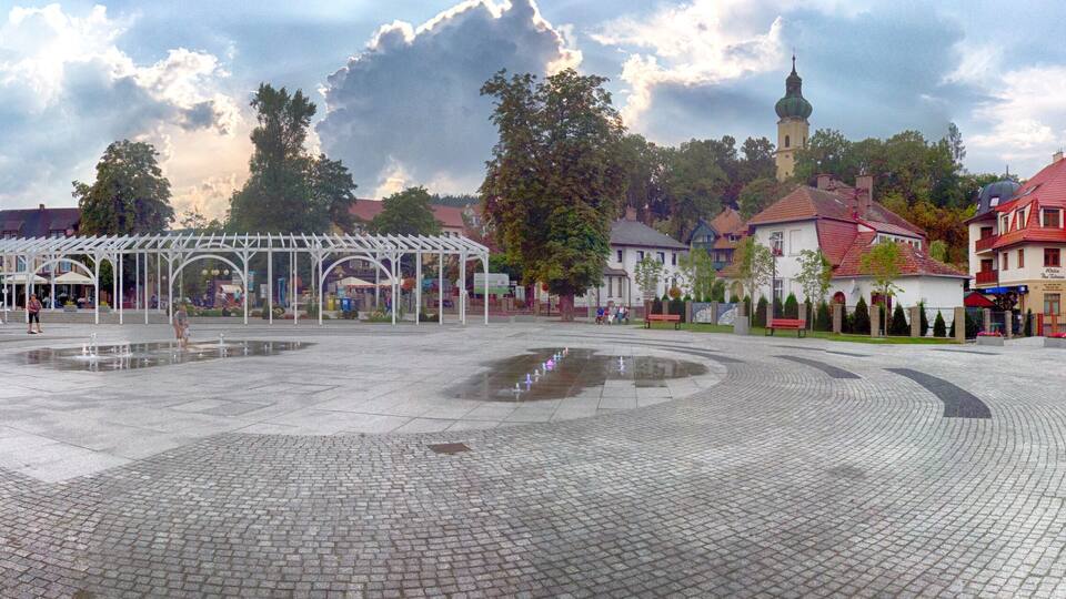 A cobblestone plaza with a white gazebo, surrounded by traditional buildings and a church with a tower.