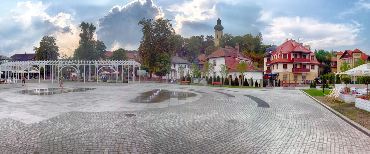 A cobblestone plaza with a white gazebo, surrounded by traditional buildings and a church with a tower.