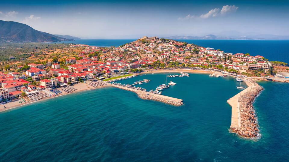 An aerial view of a coastal town with a marina, red-roofed buildings, and a mountainous backdrop.