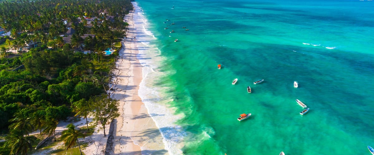 Een tropisch strand met helder turquoise water, palmbomen en kleine bootjes.
