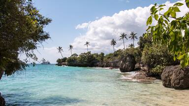 A tropical beach with clear turquoise water, palm trees, and rocky outcrops.