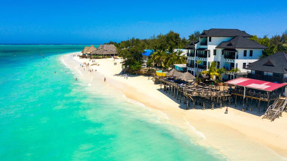 A beach with clear turquoise waters, a wooden pier, and a large white building with a red roof.