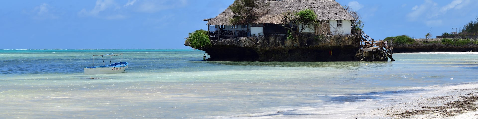 A beach with a thatched-roof house on stilts over the water, clear turquoise sea, and white sand.