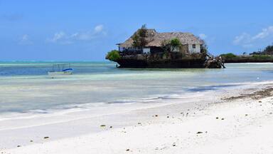A beach with a thatched-roof house on stilts over the water, clear turquoise sea, and white sand.