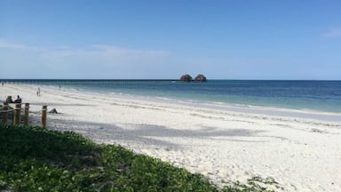 Uma praia com areia branca, um cais que se estende para o oceano e bangalôs sobre a água ao longe.