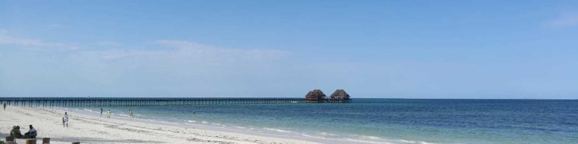 Uma praia com areia branca, um cais que se estende para o oceano e bangalôs sobre a água ao longe.