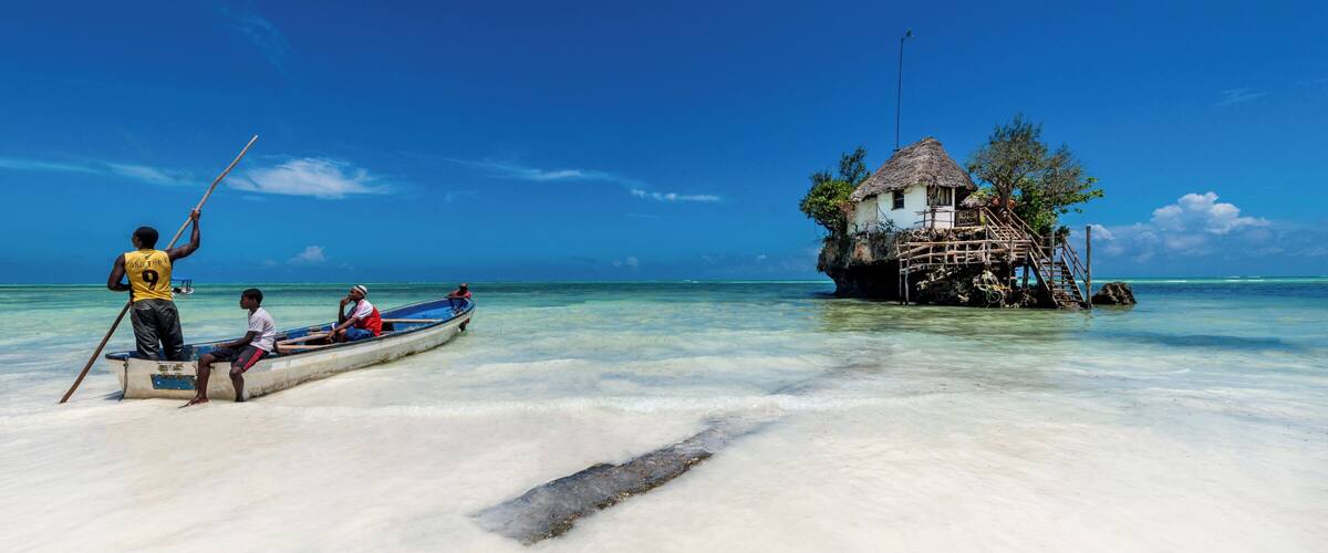 A beach with a thatched-roof house on stilts, clear turquoise water, and a boat with people.