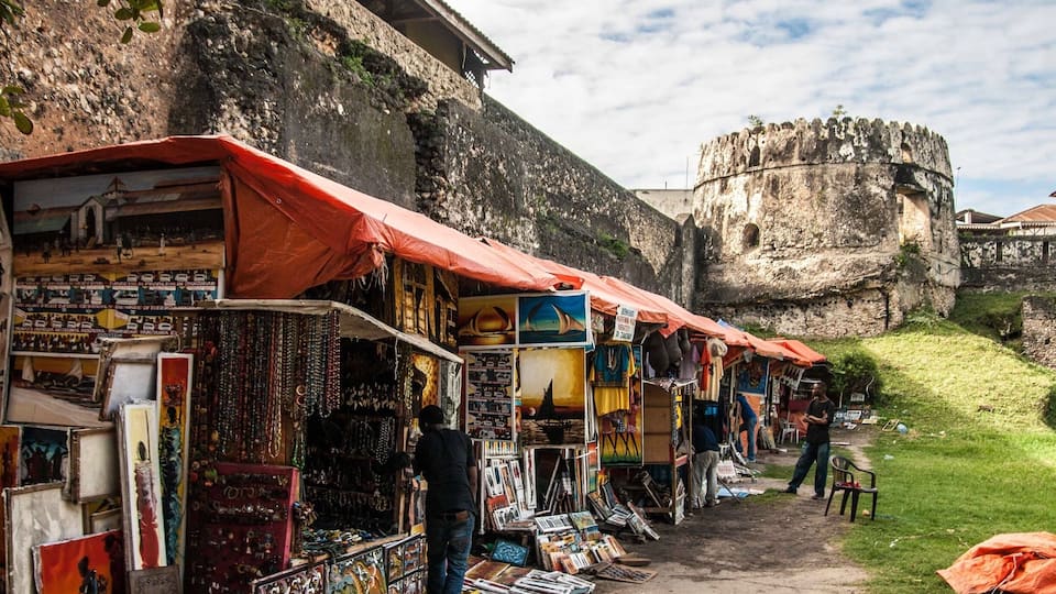 A street market with colorful stalls selling crafts and books, set against a historic stone wall and a large fortress in the background.