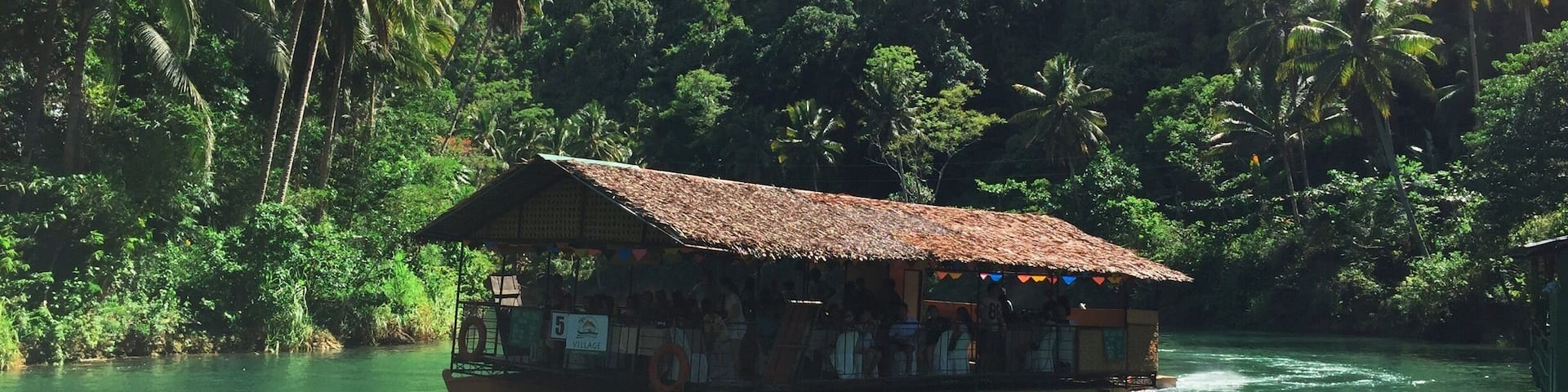 A boat with a thatched roof docked near a river, surrounded by lush greenery and palm trees.
