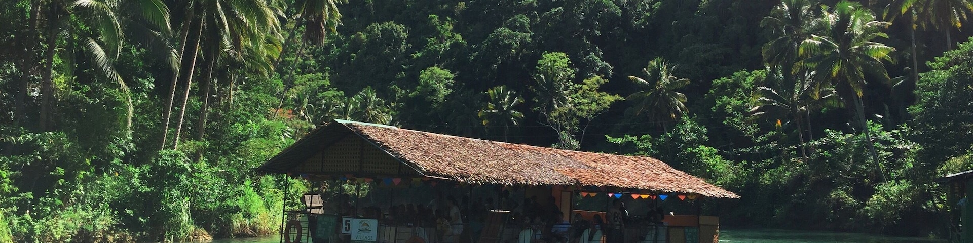 Un bote con techo de paja amarrado cerca de un río, rodeado de vegetación exuberante y palmeras.