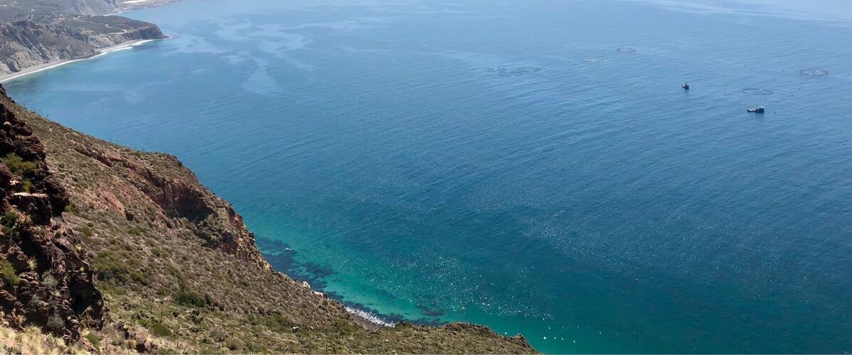 A coastal landscape with a clear blue sea, a winding road along the coastline, and distant mountains.