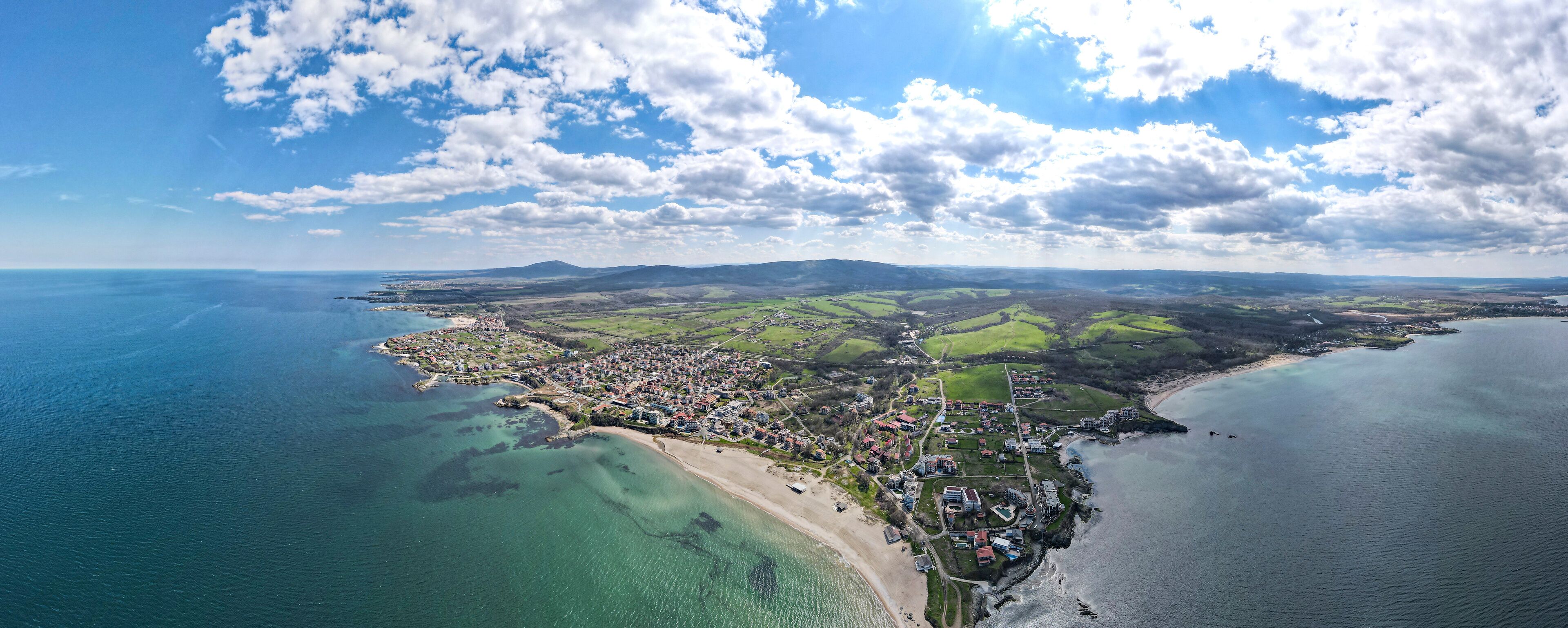 An aerial view of a coastal town with a beach, green fields, and a clear sky.