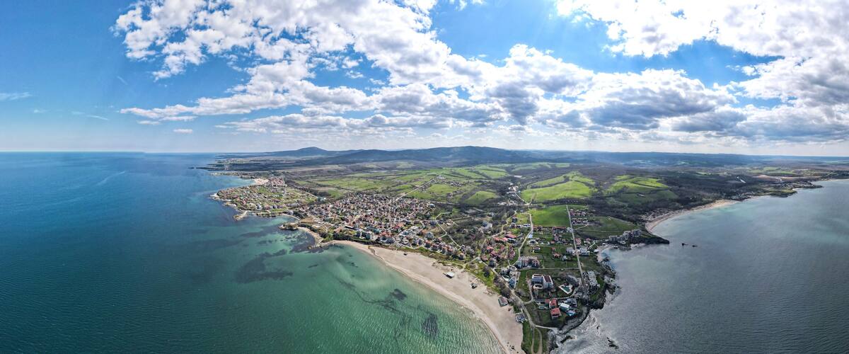 An aerial view of a coastal town with a beach, green fields, and a clear sky.
