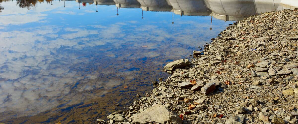 A dam with its reflection in calm water, a rocky shoreline, and autumn trees in the background.