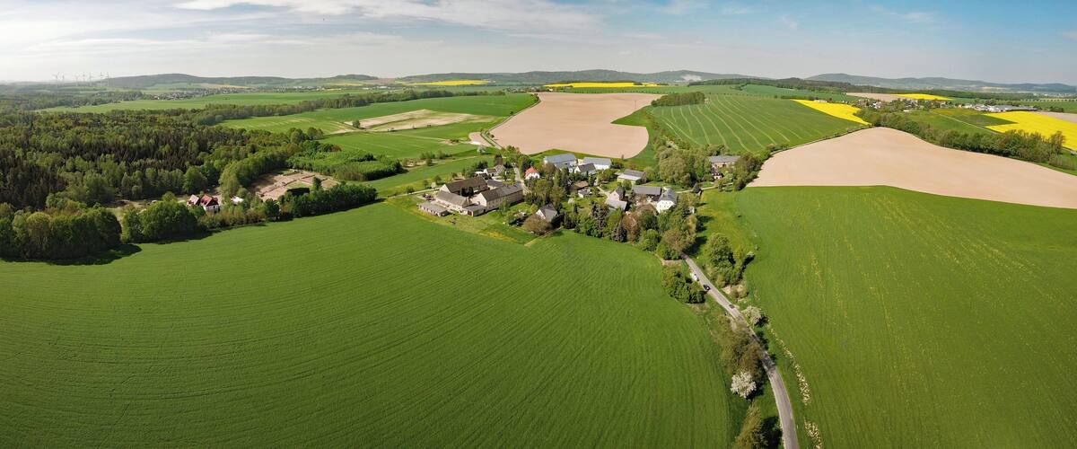 An aerial view of a rural landscape with green fields, a small village, and wind turbines in the distance.