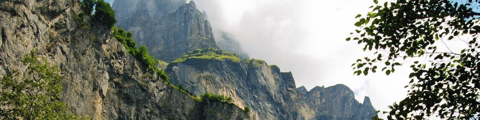 A rocky path surrounded by dense green foliage and towering mountains under a partly cloudy sky.
