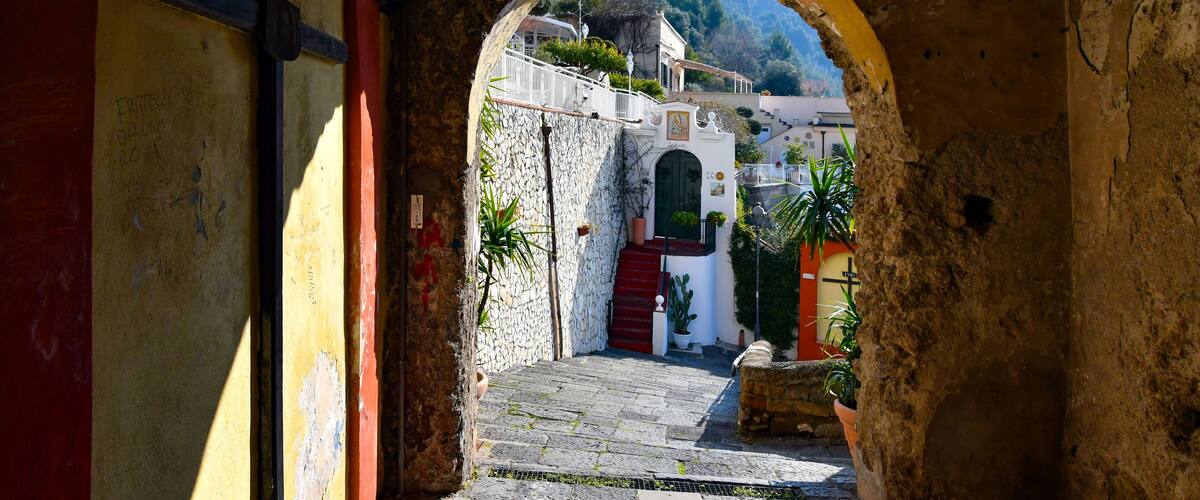 An arched stone doorway leading to a cobblestone street with a white building and red door in the background.