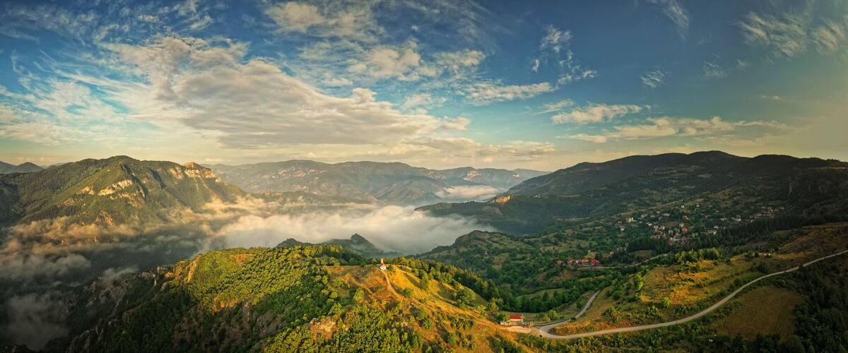 A mountainous landscape with a winding road, a small white building, and a village in the valley below.