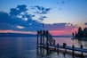 Une jetée en bois s’étendant sur des eaux calmes, surmontée d’un drapeau, entourée d’une rive boisée et baignée par la lumière d’un coucher de soleil.