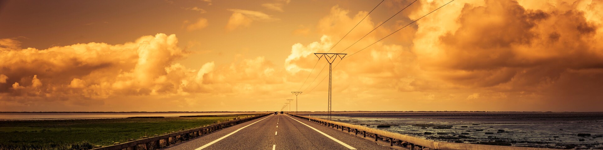 Un largo puente con barandilla, líneas eléctricas y un cielo nublado sobre un terreno llano.
