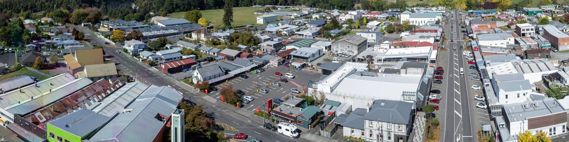 Aerial view of the town of Taihape, New Zealand, on a sunny day. The image shows the town's main street, clock tower, and surrounding buildings. Cars are driving on the road.