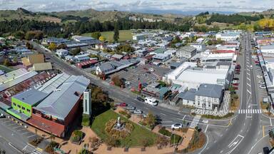 Aerial view of the town of Taihape, New Zealand, on a sunny day. The image shows the town's main street, clock tower, and surrounding buildings. Cars are driving on the road.