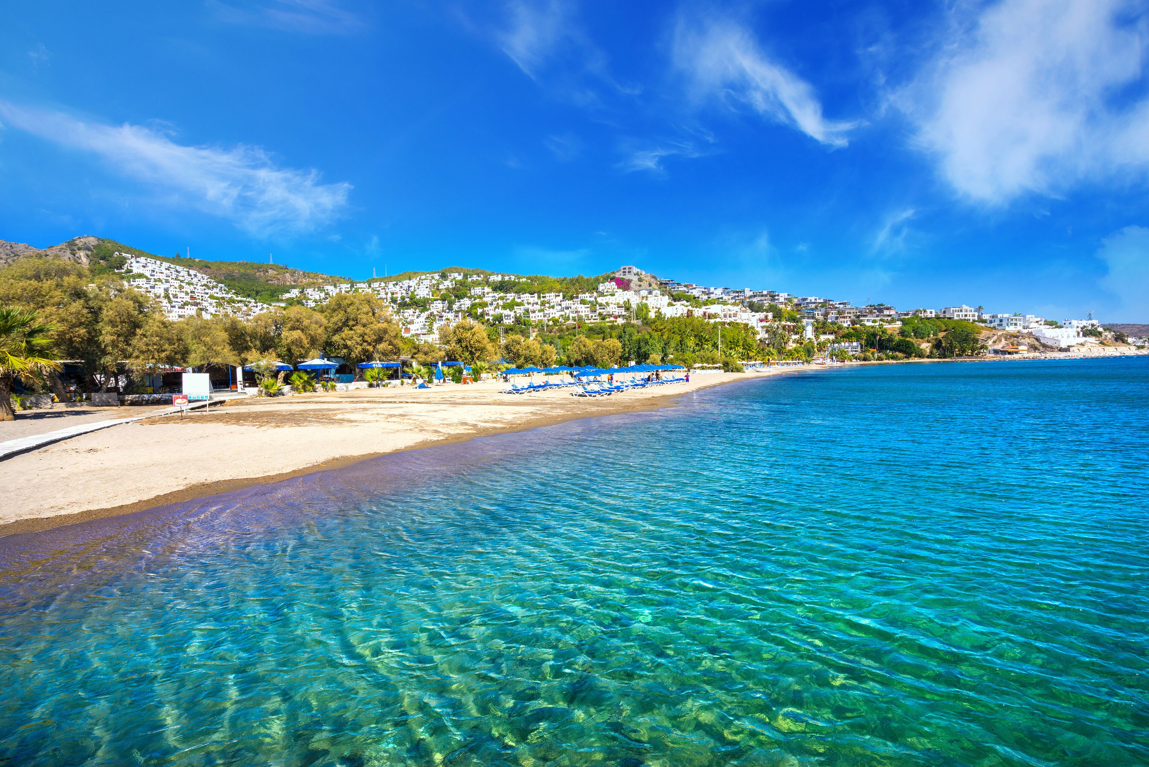 A clear blue sea, a sandy beach, and a hillside with buildings.