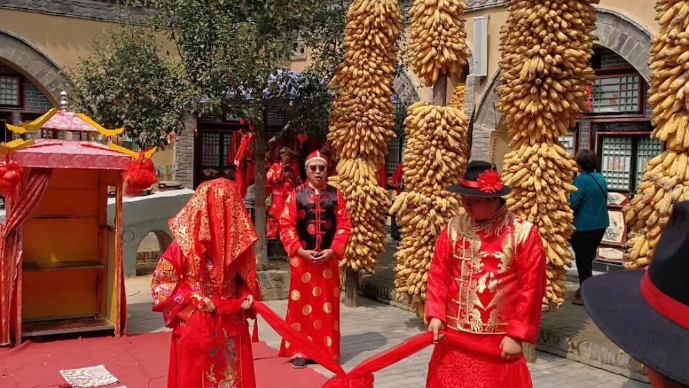 A traditional ceremony with participants in red attire, a red carpet, and a backdrop of a stone building with arches.