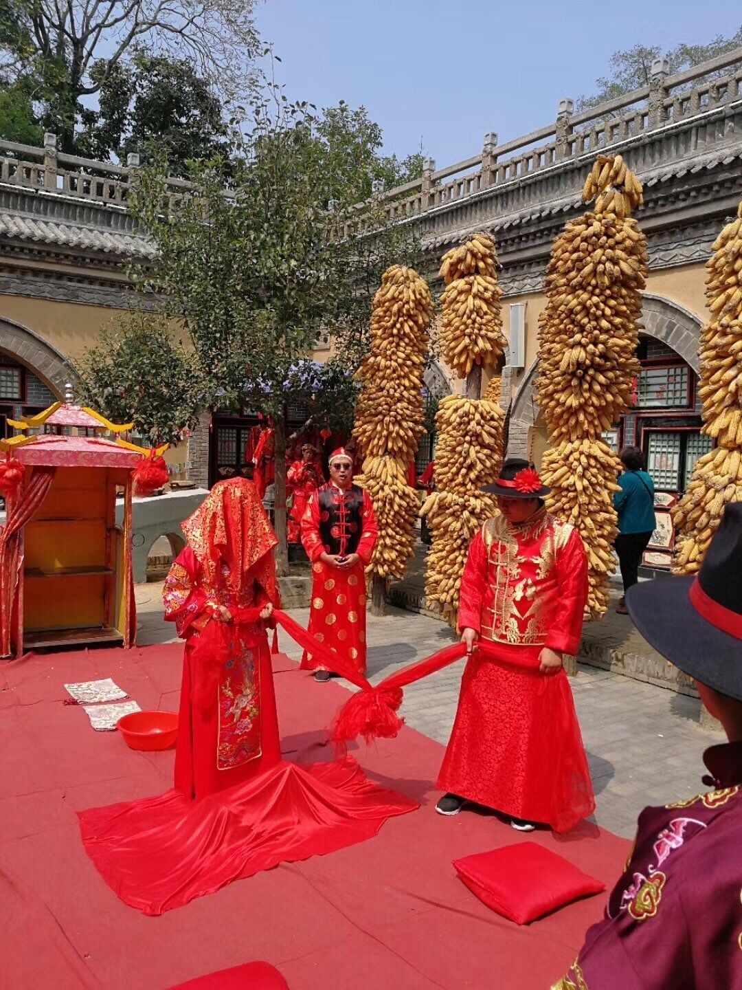 A traditional ceremony with participants in red attire, a red carpet, and a backdrop of a stone building with arches.