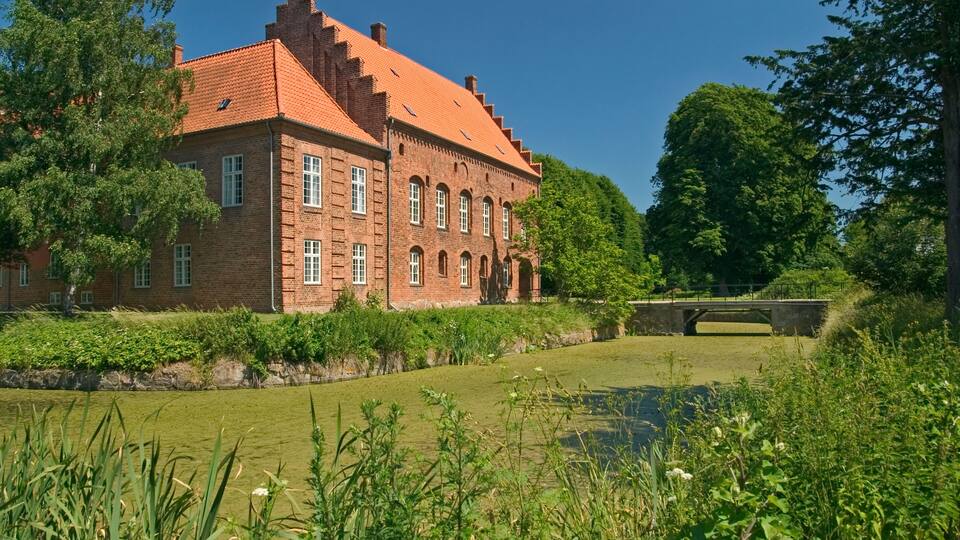 A red brick building with a tiled roof, surrounded by greenery and a canal.