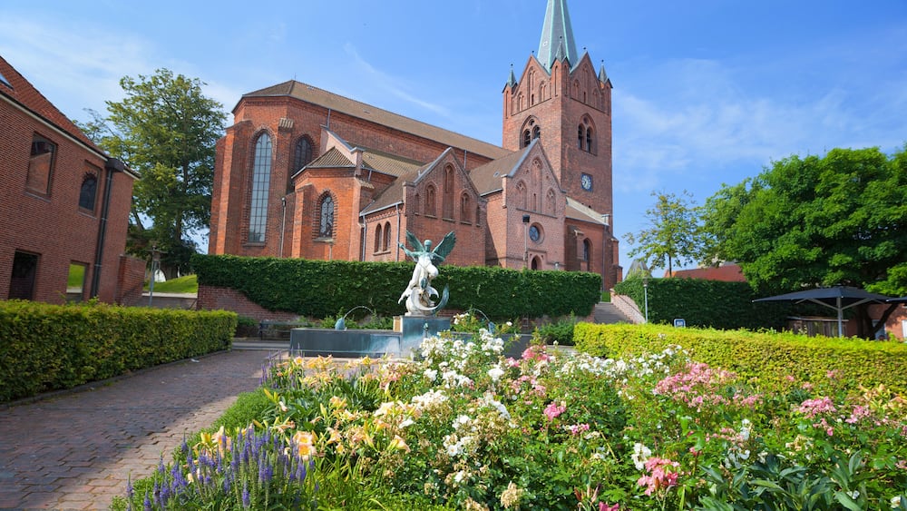 Eine historische Kirche mit einem hohen Turm, umgeben von einem gepflegten Garten mit verschiedenen Blumen und einer Statue.